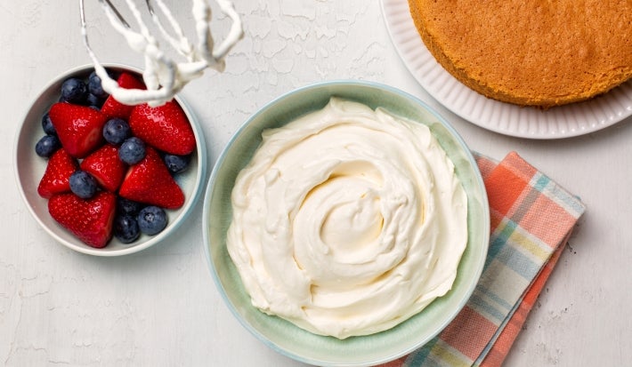 Fotografia em tons claros vista de cima de um recipiente claro fundo com creme de ninho apoiado em um pano colorido dobrado ao lado de um bolo inteiro de massa branca e um recipiente pequeno fundo com frutas vermelhas, sobre uma mesa branca.