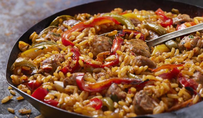 Fotografia de uma panela preta com bordas finas, com arroz amarelo, pimentão nas cores verde, amarelo e vermelho e linguiça em pedaços. No centro da panela, uma colher prata apoiada.