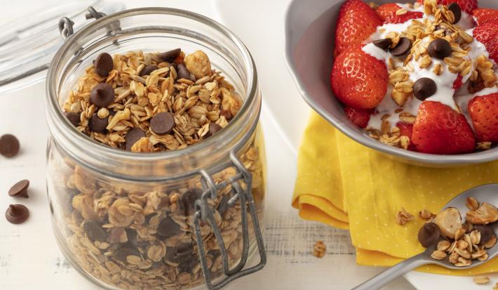 Fotografia em tons de branco de uma bancada branca com um pote de vidro com tampa  e granola. Ao lado um prato branco com paninho amarelo e uma salada de frutas com granola.