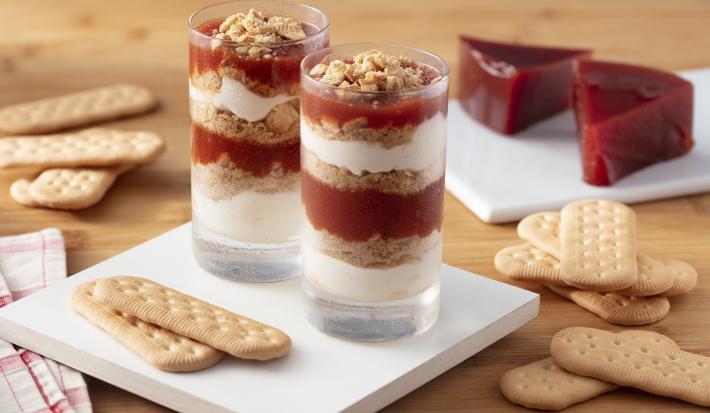 Fotografia em tons de marrom e branco de uma bancada de madeira marrom com um apoio de mesa branco quadrado, sobre ele dois copos de vidro com o verrine e biscoitos. Ao fundo biscoitos e fatias de goiabada.