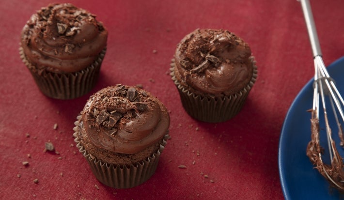 Fotografia em tons escuros de três cupcakes de nescau extra cacau com cobertura sobre um pano vermelho escuro. Ao lado tem um batedor de arame apoiado em um prato azul de vidro, raso.