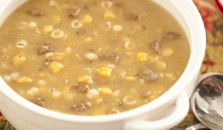 Fotografia em tons de branco e marrom de uma bancada de madeira com paninho marrom com detalhes  vermelhos, sobre ele um recipiente redondo branco com a sopa de grão-de-bico. Ao lado uma colher e ao fundo dois potes redondos vermelhos.