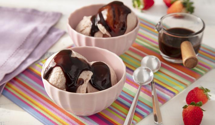 Foto da receita de Sorvete de Morango Caseiro com Calda de Leite Moça e Nescau. Observa-se dois recipientes com duas bolas de sorvete e a calda de chocolate por cima. Morangos decoram a foto.