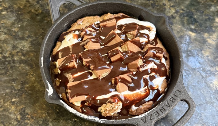 Foto vista de cima da receita de S'Mores de KitKat, preparada e servida em uma pequena panela de ferro, decorada com muita calda de chocolate e pedaços de KitKat