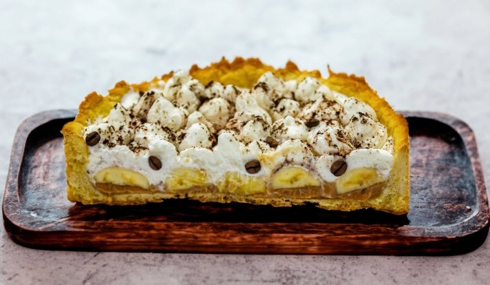 Em cima de uma mesa, banoffee cortada ao meio com massa de biscoitos crocante, bananas em rodelas, doce de leite, decorado com chantilly e canela em pó.