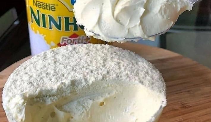 Foto da receita de Mousse de Leite Ninho Simples. Observa-se uma travessa grande de vidro com o mousse dentro e uma colher bem perto expondo a cremosidade da receita.