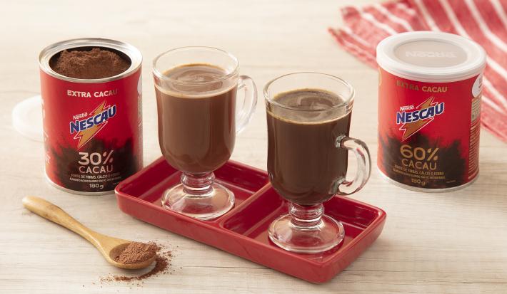 Receita do cappuccino thermogenico feita com NESCAU Extra Cacau servida em duas xícaras de vidro sobre um suporte de porcelana vermelho. Esses elementos sobre uma mesa de madeira com duas latas de nescau, um pano listrado e uma colher de nescau decorando.