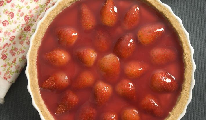 Fotografia em tons de vermelho com em uma bancada de madeira clara com um pano vermelho, um prato branco raso grande com a torta de morango.
