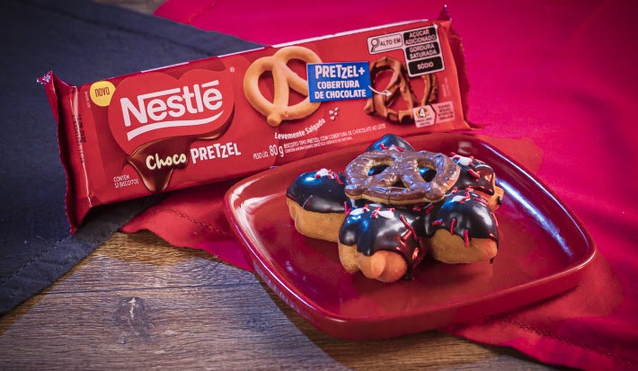 Donut em formato de flor, coberto com chocolate escuro e branco, detalhes vermelhos e um ChocoPretzel crocante no centro.