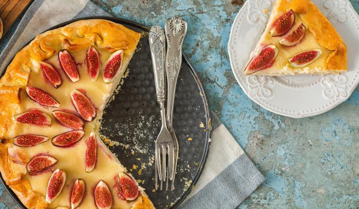 Fotografia vista de cima de uma torta de amêndoas com creme de leite e figos, coberta com figos fatiados, sobre o fundo falso da forma e uma fatia ao lado