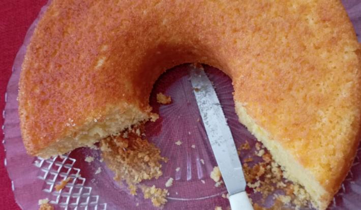 Fotografia vista de cima de um bolo de fubá quase inteiro sobre um prato raso de vidro grande sobre uma toalha de mesa vermelha.