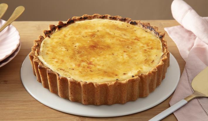 Foto da receit ade torta de limão brulée servida em uma porção grande sobre um prato branco em cima de uma mesa de madeira com um paninho rosa ao lado