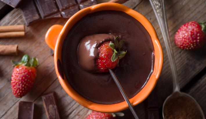 Fotografia mostra um morango mergulhado em uma panela laranja com chocolate derretido.