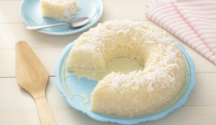 Fotografia em tons de azul e rosa de uma bancada de madeira branca, ao centro um prato azul com o cuscuz de tapioca coberto com coco ralado. Ao lado uma espátula de madeira em um pano com listras rosas e brancas.