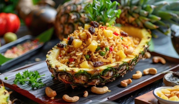 Em um abacaxi cortado ao meio, porção de arroz com abacaxi picados, ervilhas, uvas-passas e castanha-do-Pará.