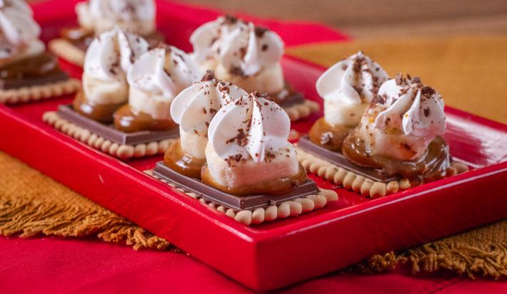 Fotografia em tons de vermelho com vários biscoitos Choco Biscuit ao centro. Por cima de cada biscoito existe uma camada de doce de leite, banana e, por cima, chantilly.
