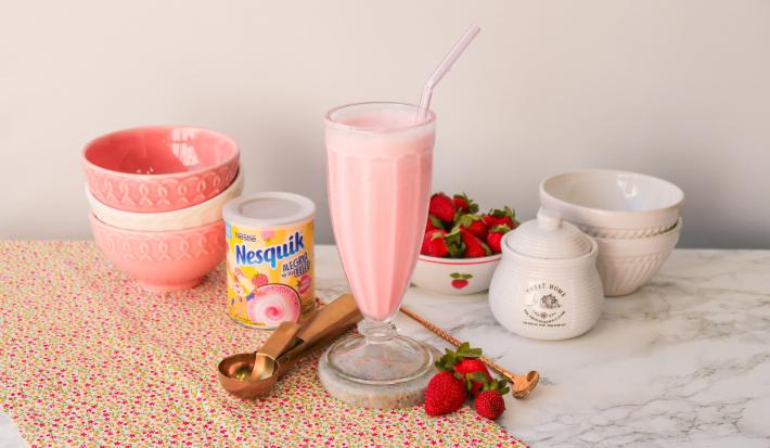 Fotografia de um milkshake rosa servido em uma copo alto de vidro. Ao fundo um cenário com morangos e bowls com tons rosas.