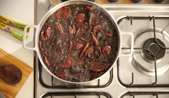 Foto vista de cima de um fogão e uma bancada. Sobre o fogão há uma panela branca com a receita de feijoada dentro, com uma fumaça saindo.