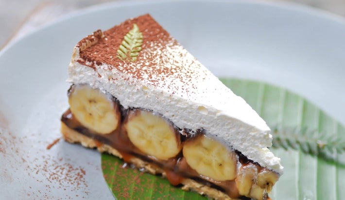 Em cima de uma mesa, prato com fatia de banoffee com massa crocante, doce de leite cremoso e bananas em rodelas, decorado com chantilly e chocolate em pó.