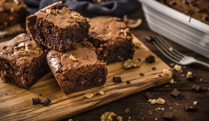 Fotografia de quatro pedaços de brownie feito de farinha de soja, castanha e chocolate. Os pedaços estão sobre uma tábua de madeira perto de um garfo.