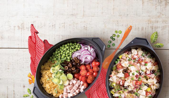 Fotografia em tons de cinza, vermelho, preto de uma bancada cinza vista de cima, contém um pano vermelho e por cima dois recipientes ovais um com macarrão, azeitona, peito de peru e tomates picados e no outro os mesmos ingredientes só que inteiros.