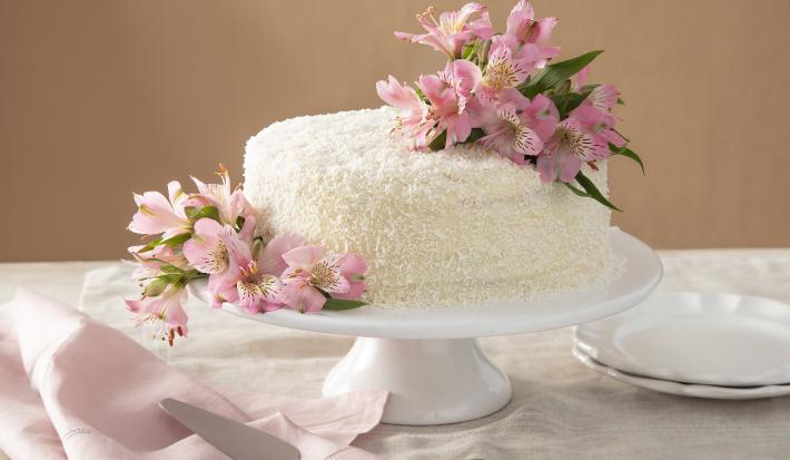 Foto da receita de Bolo de Gala. Observa-se um bolo grandioso com flores rosas sobre uma boleira de cerâmica branca. Atrás, um fundo nude e pratinhos de sobremesa decoram a foto, assim como um guardanapo rosa.