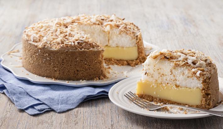 Fotografia em tons de marrom e azul de uma bancada de madeira com um paninho azul, sobre ele um prato branco com uma torta de coco. Ao lado um prato pequeno redondo com uma fatia da torta e um garfo.