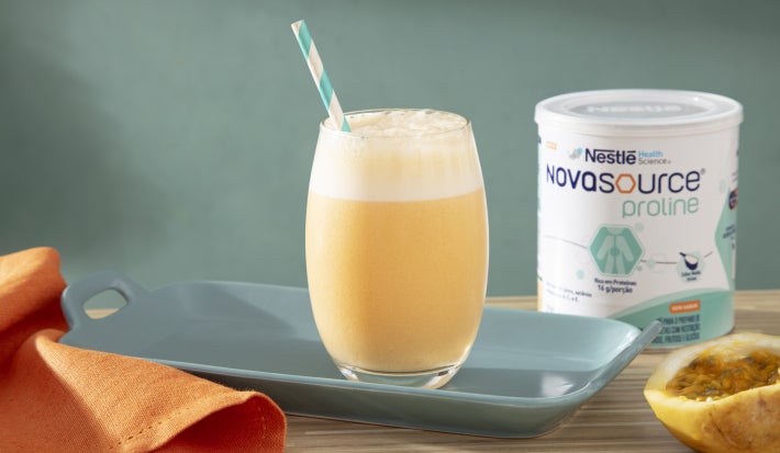 Foto da receita de vitamina de maracujá servida em um copo largo de vidro sobre uma louça de porcelana azul em cima de uma mesa de madeira com uma embalagem de novasource proline ao lado