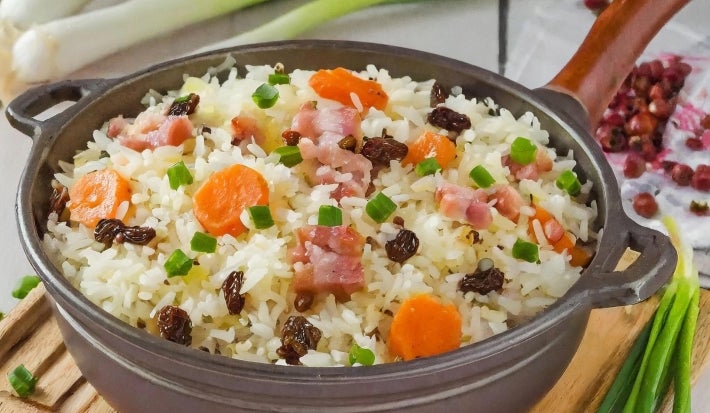 Em um recipiente redondo cinza de porcelana, há uma base de arroz soltinho, combinado com pedaços de presunto cozido, coloridos vegetais, como cenoura, uvas-passas e decorado com cebolinha picada.