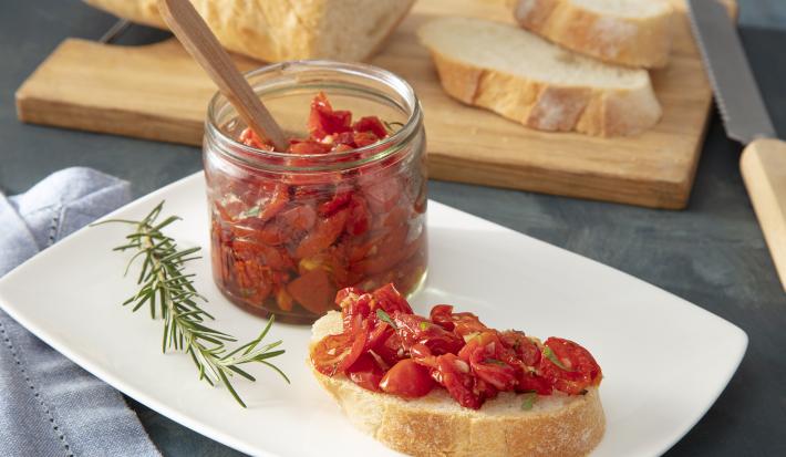 Foto da receita de Tomate confit. Observa-se uma torrada com os tomates assados em cima e um recipiente transparente de vidro com os tomates armazenados. Alecrim e pedaços de pão fatiados decoram a foto.