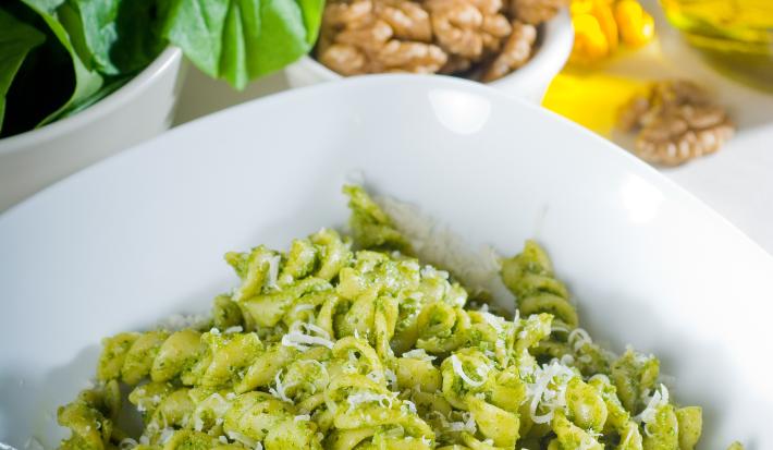 Fotografia em tom claro e branco, com um prato quadrado fundo branco no centro da foto, dentro tem macarrão no estilo parafuso com pesto em tom verde claro e queijo ralado por cima. No fundo da foto tem manjericão e pinolis, tudo sobre uma mesa branca..