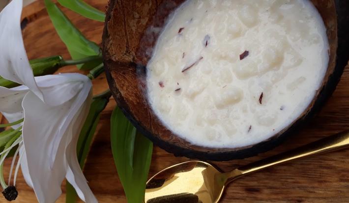 Foto da receita de canjica de coco fresco servida em um pote de vidro sobre uma mesa de madeira com uma colher dourada e um flor branca ao lado