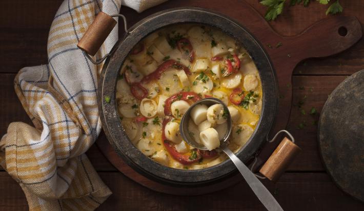 Fotografia em tons de marrom e grafite e branco, com panela de pedra ao centro com moqueca de palmito e uma concha, sobre tábua de cozinha, no entorno tampa, ramos de salsa e guardanapo colorido listrado, tudo sobre bancada de madeira.