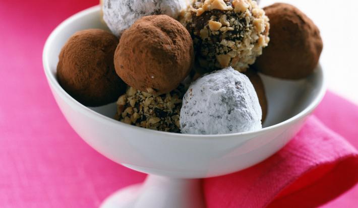Fotografia em tom claro, um pote branco decorativo sobre uma toalha de mesa rosa. Dentro desse recipiente, tem várias trufas de chocolate, algumas enroladas em nozes trituradas, outras em cacau e três delas passadas em açúcar de confeiteiro.