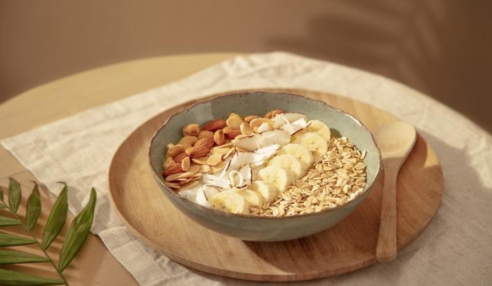 Foto em tons de bege da receita de bowl de manga com aveia servida em um bowl de cerâmica irregular sobre uma tábua circular de madeira com uma colher de madeira ao lado. Tudo sobre uma mesa com uma toalinha bege mais clara por cima