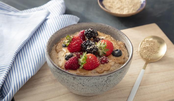 Foto em tons de bege da receita de mingau nutritivo servida em um bowl de cerâmica sobre uma tábua de madeira clara com uma colher prateada ao lado. Ao lado ainda há um paninho azul listrado e um recipiente com aveia em flocos
