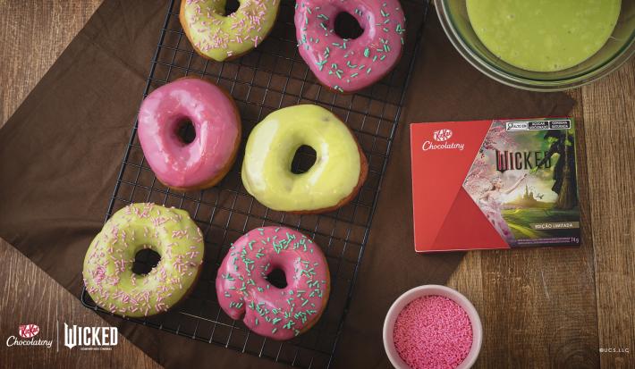Foto da receita de Donuts Wicked, vistos de cima, servidos em uma grade de ferro preta. São seis no total, decorados com cobertura rosa e verde e alguns com granulados coloridos. Ao lado direito há uma caixa de KitKat Wicked.