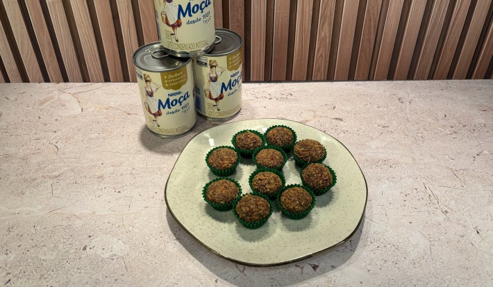 Prato com docinhos cobertos com crocante em forminhas verdes e latas de Leite Moça ao fundo.