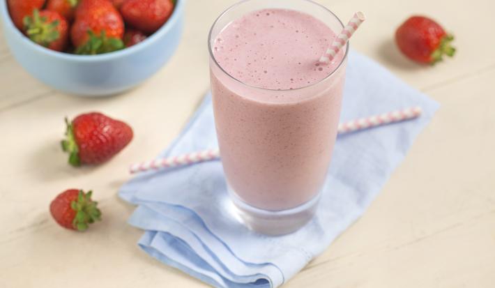 Fotografia em tons de rosa e azul de uma bancada rosa com um paninho azul, sobre ele um copo de vitamina com um canudo. Ao lado morangos. Ao fundo um recipiente redondo azul com morangos.