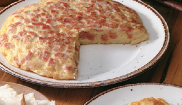 Fotografia em tons de vermelho em uma mesa de madeira com um prato branco raso grande com a omelete cortada ao meio. Ao lado, um prato com a fatia da omelete e outro prato com fatias de pão.