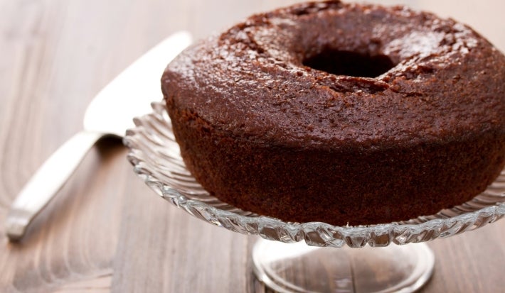 Um bolo de chocolate em forma de bundt, com cor marrom escura e superfície brilhante, está sobre um elegante suporte de vidro. O ambiente é rústico e sofisticado, sugerindo que o bolo está pronto para ser servido.