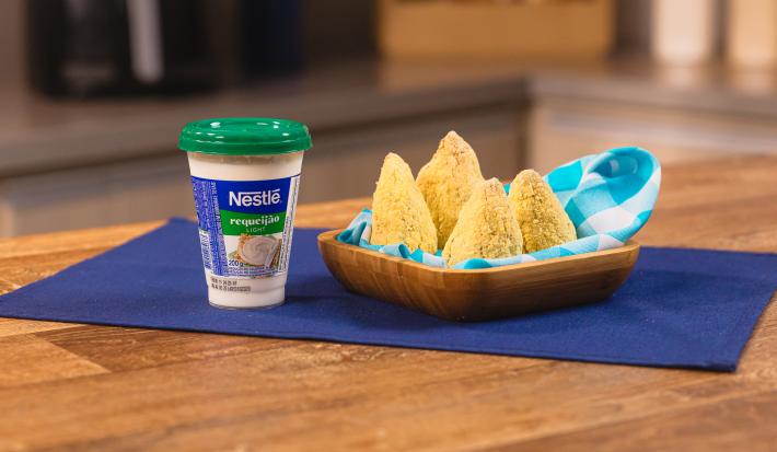 Fotografia de uma bancada de madeira com um pano azul escuro. Sobre ele tem um recipiente quadrado de madeira com um pano azul e branco forrando as quatro coxinhas saudáveis que estão dentro. Ao lado do recipiente tem um pote de requeijão light.
