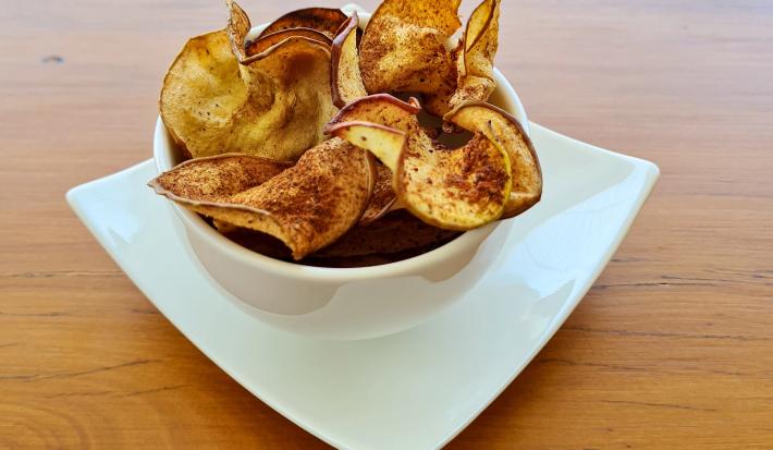 Foto da receita de Chips de Maçã. Observa-se um potinho branco de porcelana com os chips dentro polvilhados de canela.