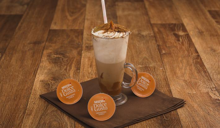 Fotografia em tons de marrom em uma bancada de madeira escura, um pano marrom, cápsulas de Mochaccino Canela Dolce Gusto e uma xícara de vidro alta com a bebida feita com banana, doce de leite e chantilly.