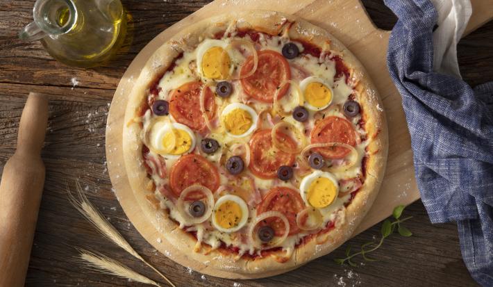 Fotografia em tons de azul em uma bancada de madeira escura, um paninho azul, uma tábua de madeira com a pizza portuguesa em cima dela. Ao lado, um potinho de azeite e um rolo de massa.