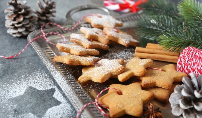Em cima de uma mesa decorada com enfeites de natal, prato com biscoitos dourados e crocantes em formato de estrela, decorados com açúcar refinado.
