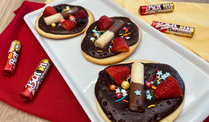 Fotografia em tons de vermelho e amarelo com um recipiente branco ao centro. Em cima do recipiente existe 3 mini pizzas doces cobertas com ganache de chocolate meio amargo, morangos e BATON Duo. Ao lado existe algumas embalagens de BATON Duo