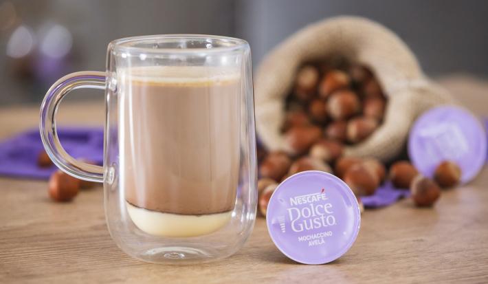 Fotografia em tons de lilás em uma bancada de madeira, uma xícara de vidro com a bebida de Mochaccino Avelã dentro, ao lado uma cápsula de Dolce Gusto, atrás um saco de juta com avelã e um paninho lilás.