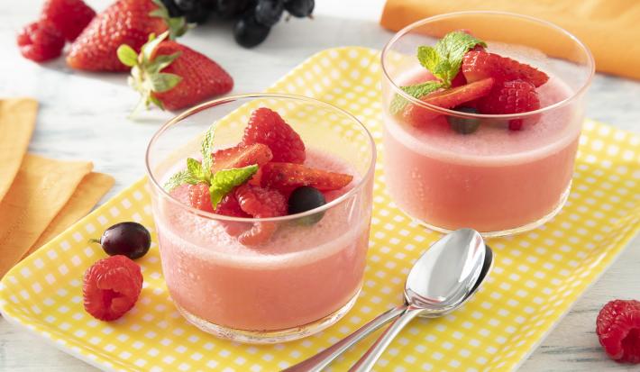 Foto da receita de Gelatina Cremosa. Observa-se dois potes de vidro com a gelatina vermelha dentro e morangos e mirtilos decorando o topo.
