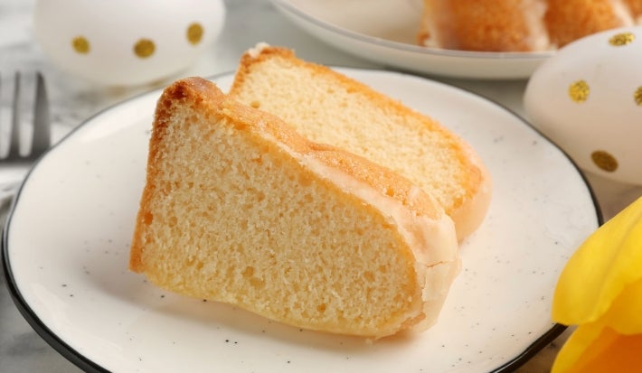 Fotografia de fatias de um bolo de queijo leve e fofinho em um prato elegante. O cenário festivo inclui ovos decorados e uma pétala de flor amarela, transmitindo uma atmosfera de celebração e aconchego.